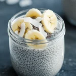 Glass jar of Coconut Banana Chia Overnight Paleo Oats topped with banana slices and coconut flakes on a dark surface.
