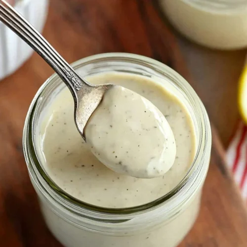 Paleo Caesar Salad Dressing: Clean, Creamy & Anchovy-Free Options 2 Hand scooping paleo Caesar salad dressing from a glass jar with a spoon, surrounded by lemons and another jar.