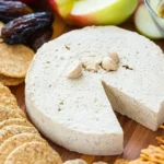 Paleo cheese wheel served on a wooden board with grain-free crackers, apple slices, dates, and pickles.