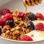 A bowl of paleo granola with raspberries, blackberries, and dairy-free yogurt being drizzled with honey.