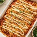 Overhead view of a baked spaghetti dairy free casserole topped with plant-based cheese and parsley in a white dish.