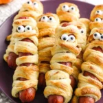 Halloween Sausage Idea – hot dogs wrapped in puff pastry like mummies with candy eyes on a purple plate.